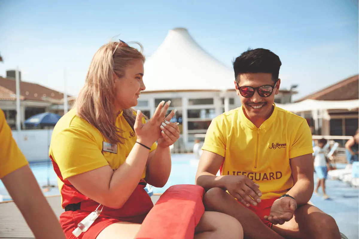 Two lifeguards in yellow and red uniforms sitting together by an outdoor pool, talking and smiling.