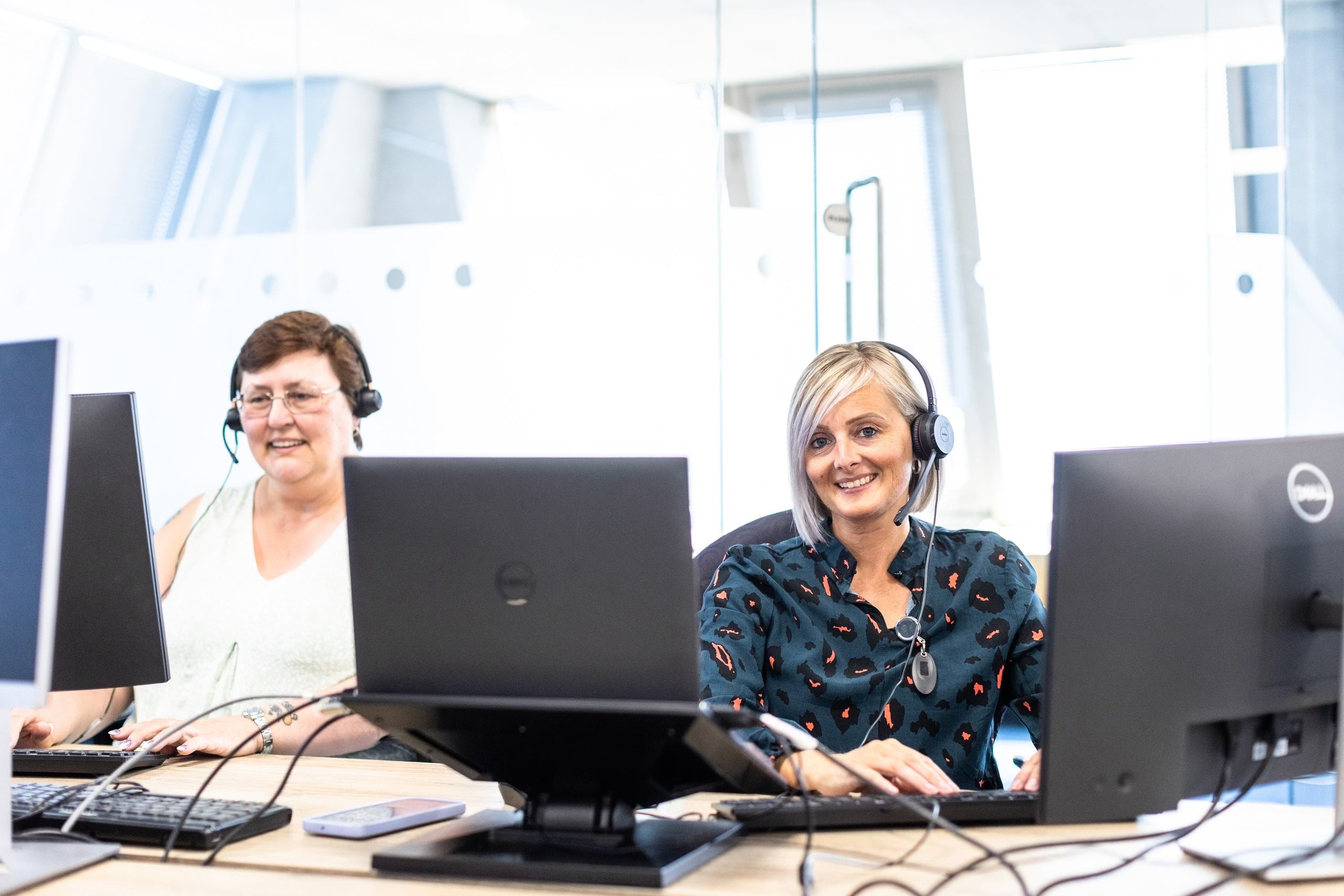Two women wearing headsets working at desks with computers in an office contact center.