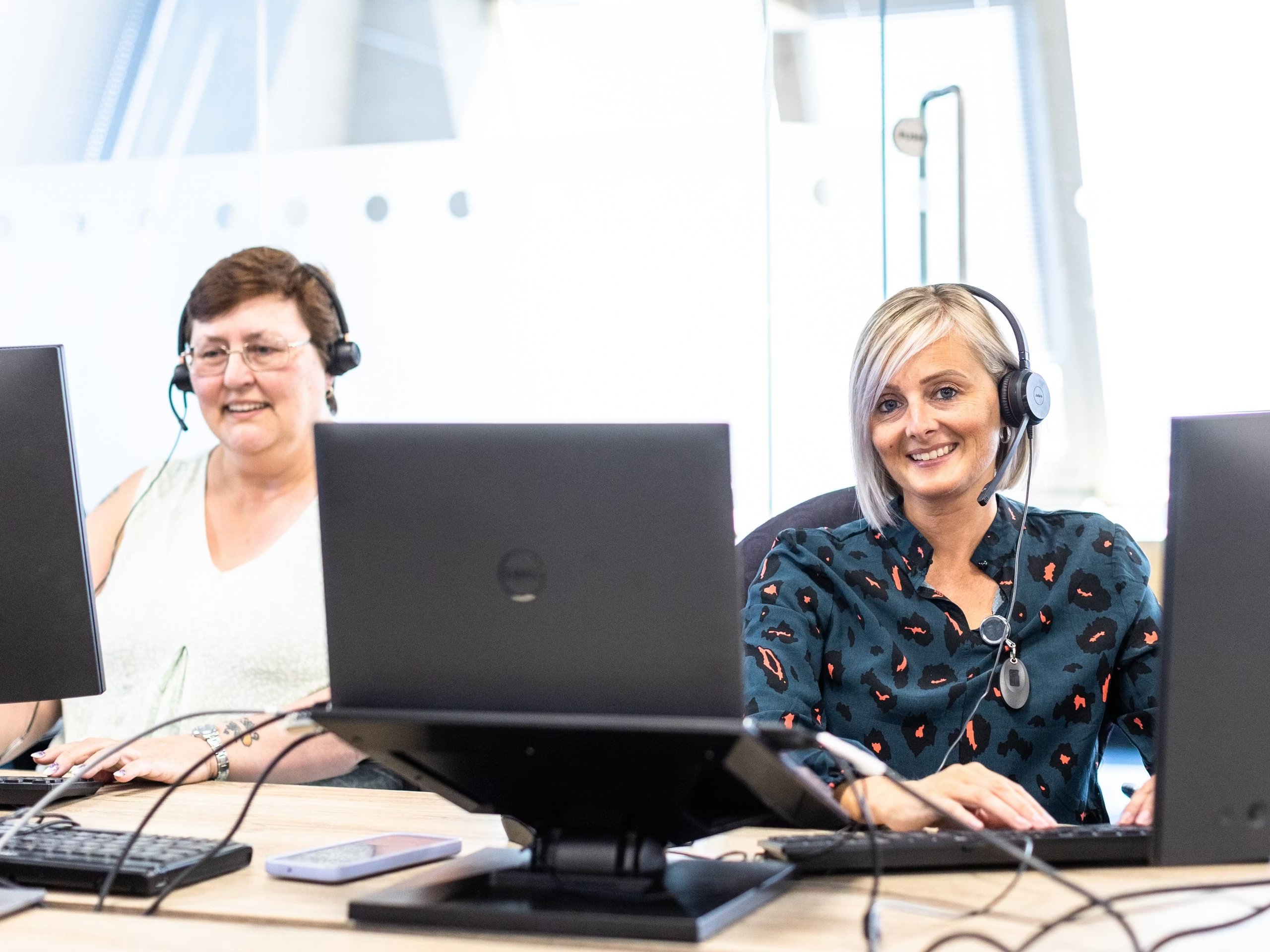 Two women wearing headsets working at desks with computers in an office contact center.