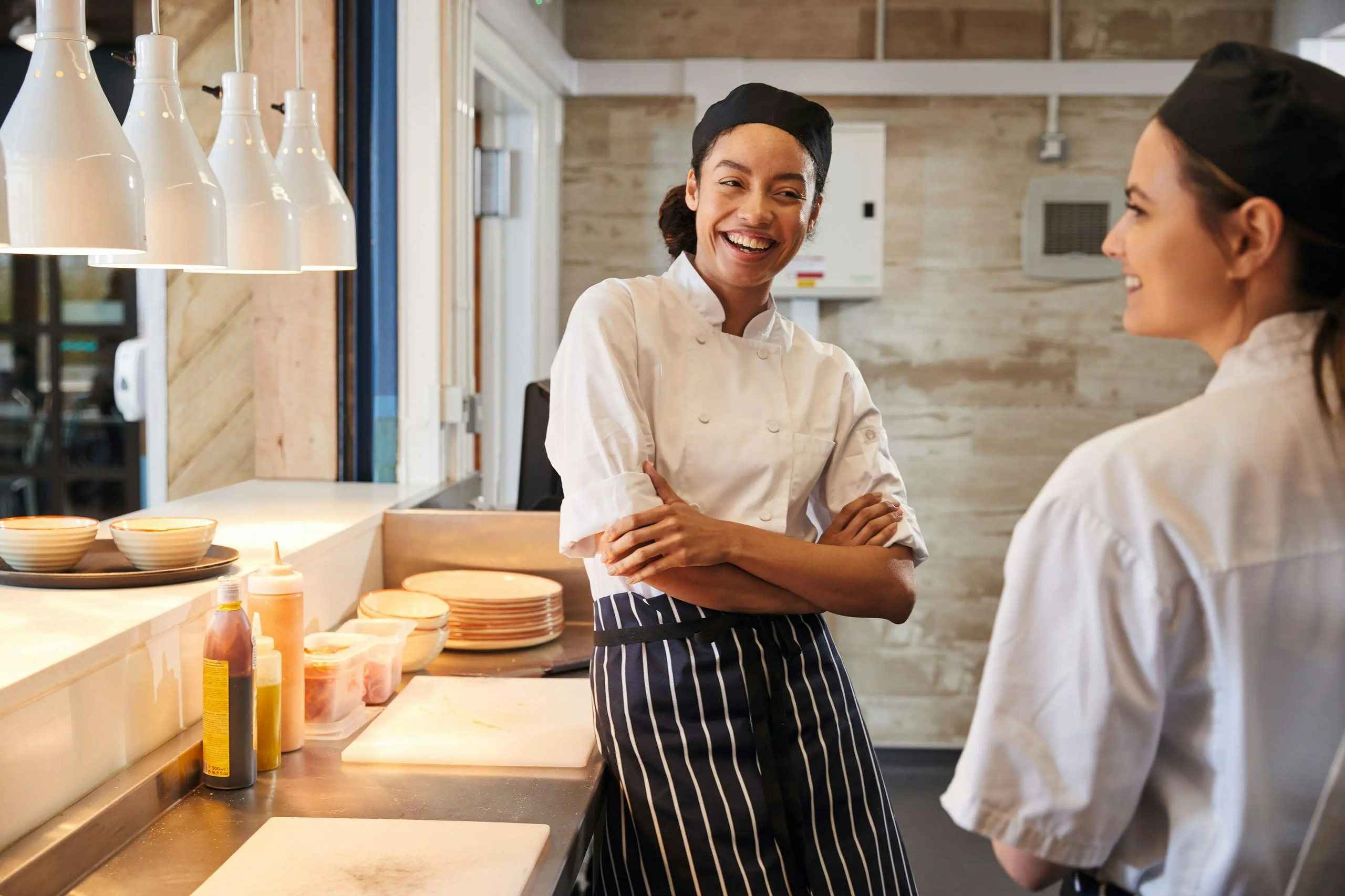 Two female chefs wearing white uniforms and black aprons laughing together in a restaurant kitchen.