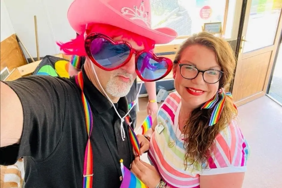 A man in a pink cowboy hat and heart sunglasses taking a selfie with a woman wearing rainbow stripes.