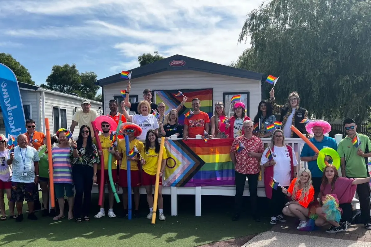 A large group of people celebrating Pride, holding rainbow flags in front of mobile homes.