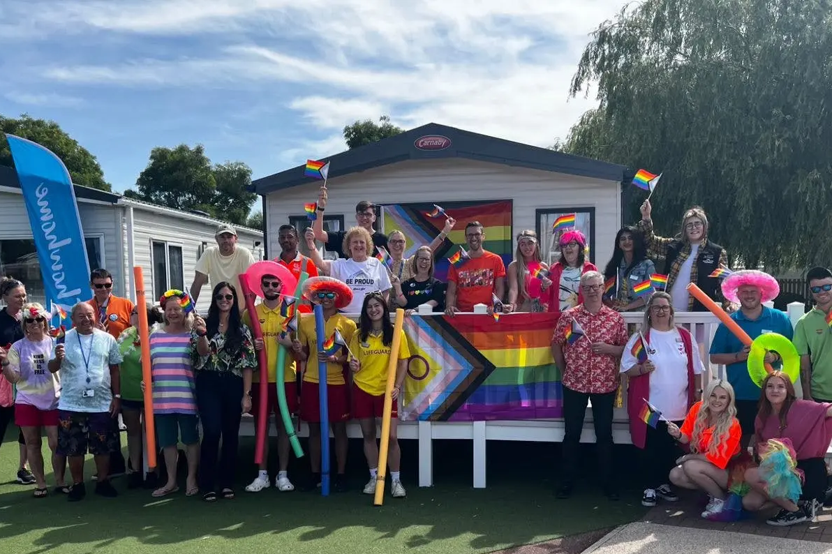 A large group of people celebrating Pride, holding rainbow flags in front of mobile homes.