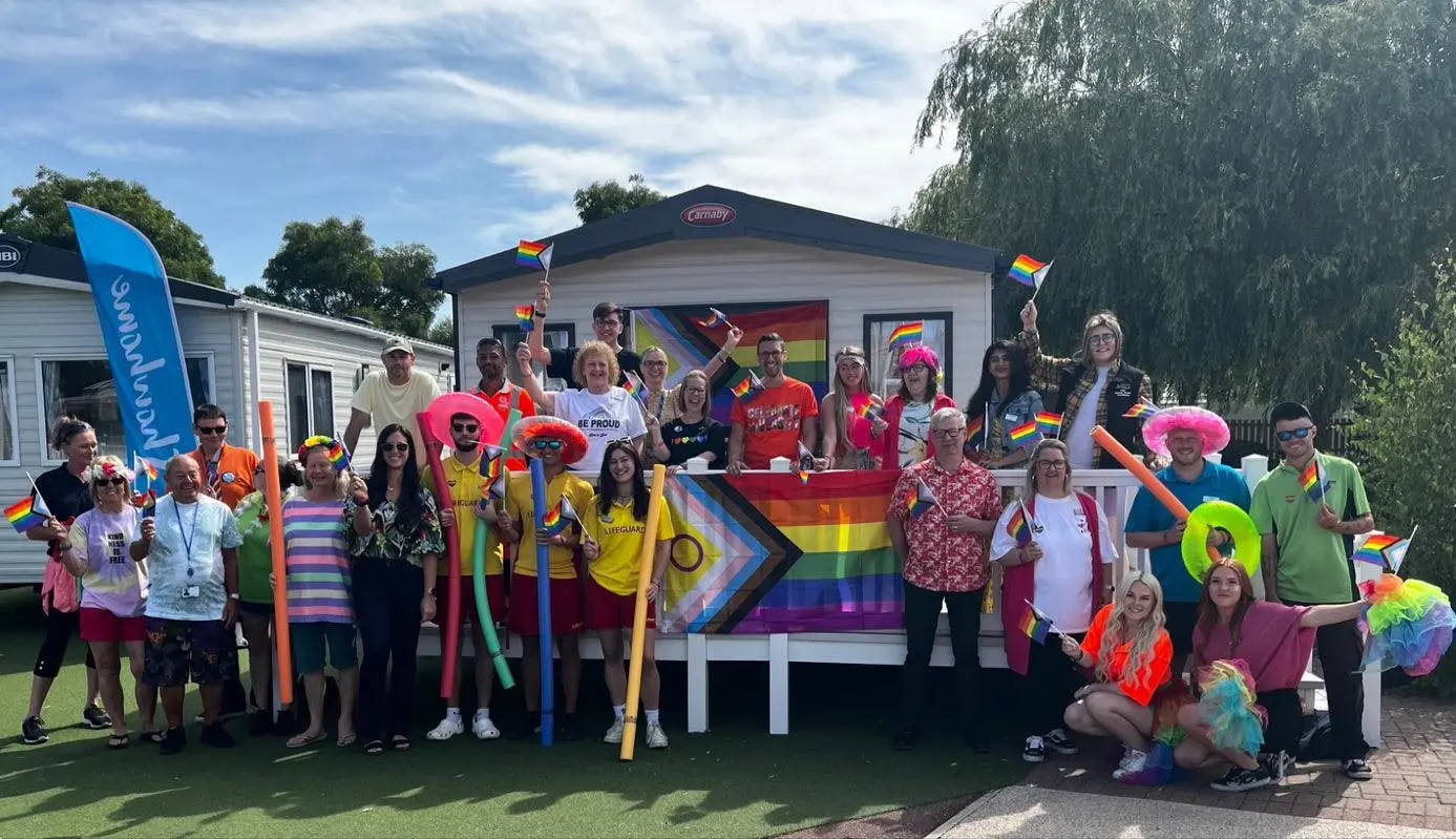 A large group of people celebrating Pride, holding rainbow flags in front of mobile homes.