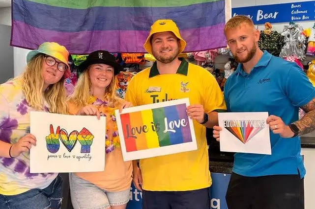 Four smiling people hold Pride signs in front of a large rainbow flag.