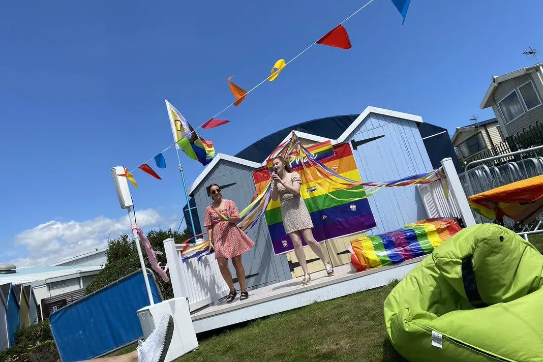 Two people on a stage decorated with rainbow flags and bunting during an outdoor Pride event.