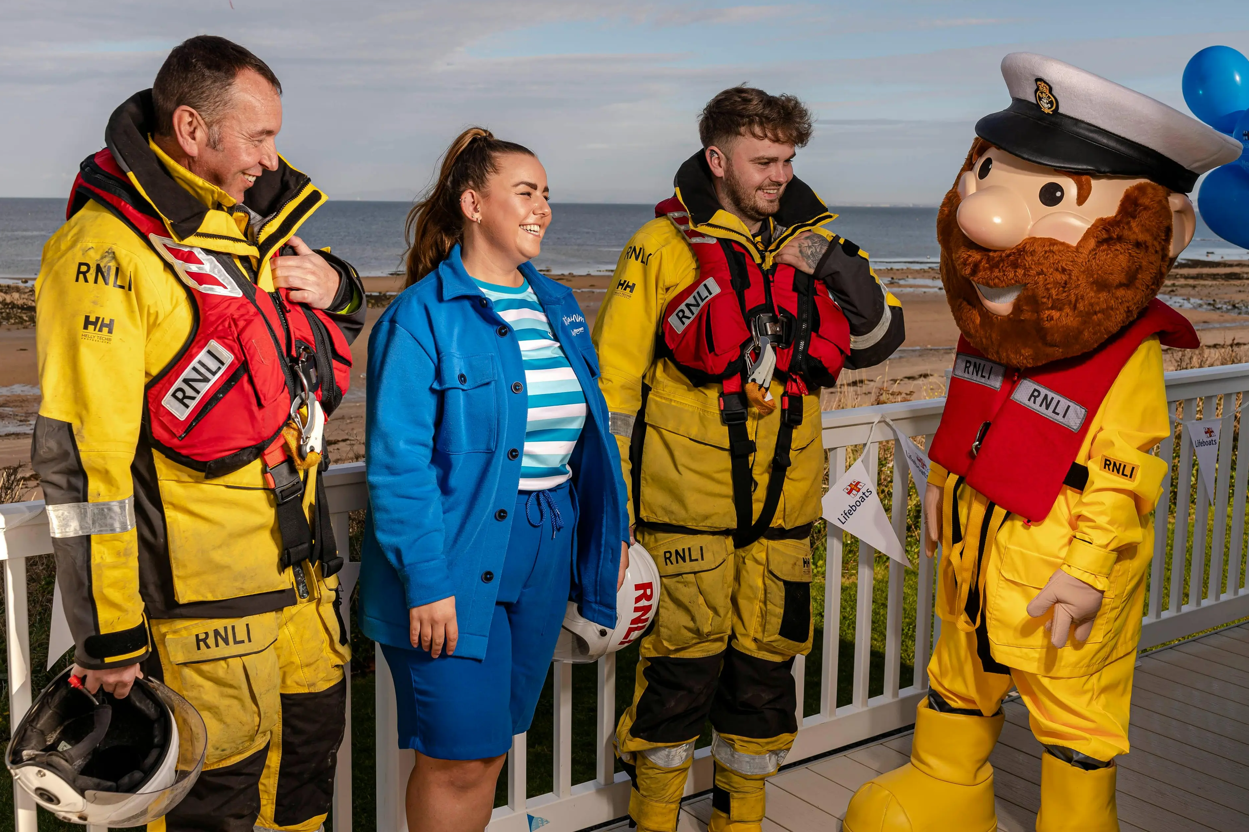 RNLI crew members in yellow gear and a woman in blue stand next to an RNLI mascot by the coast.