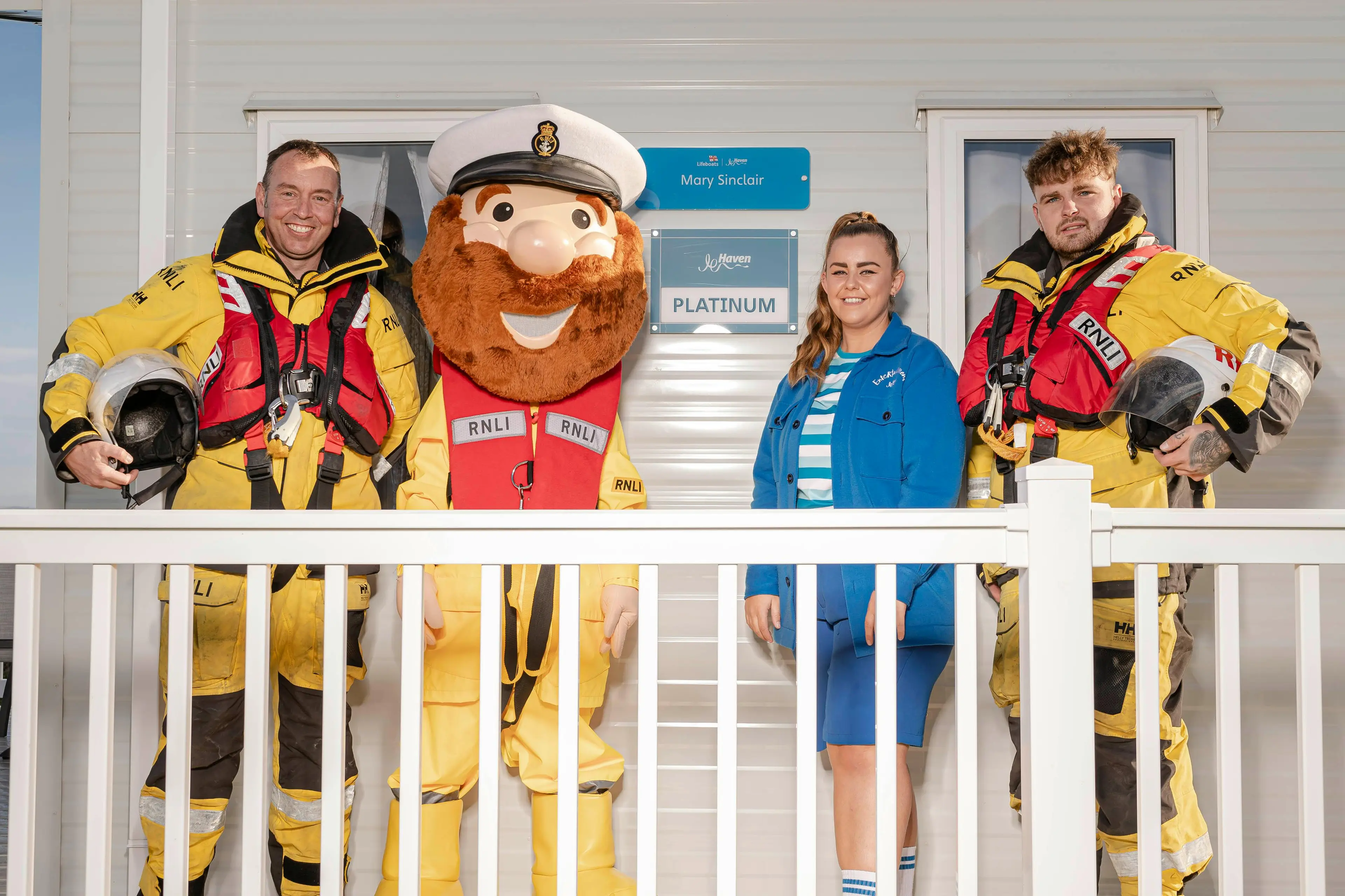 Two RNLI crew members in yellow gear, a mascot in a captain's hat, and a woman pose together.