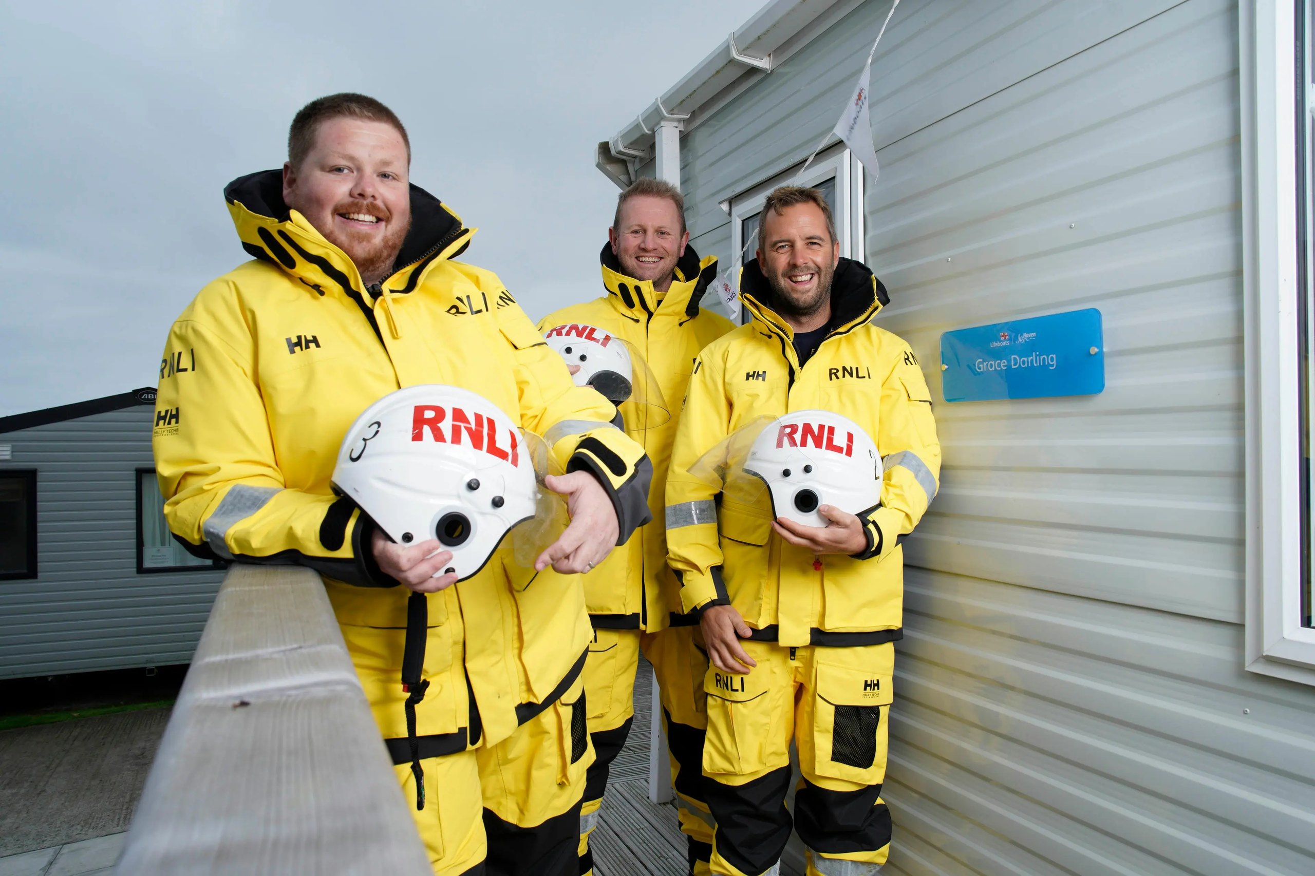 Three smiling RNLI crew members in yellow waterproof gear holding RNLI helmets outside a building.