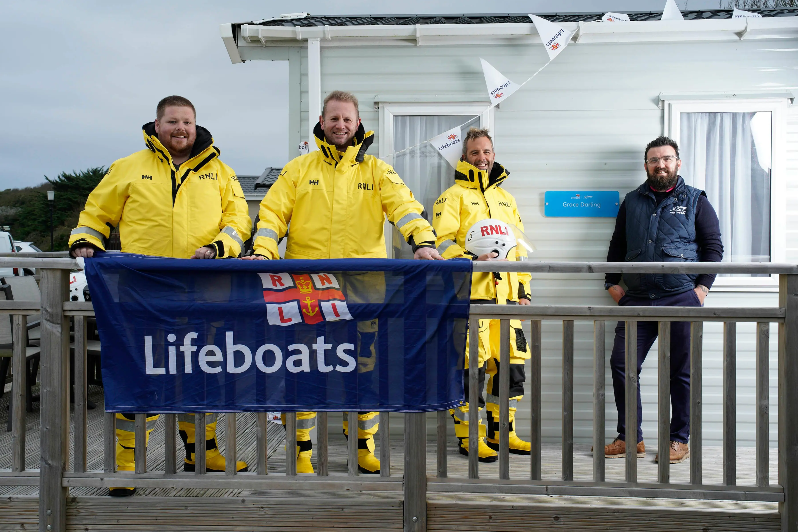Four men standing on a wooden deck in front of a white building, with three wearing yellow RNLI jackets and one holding an RNLI Lifeboats flag.