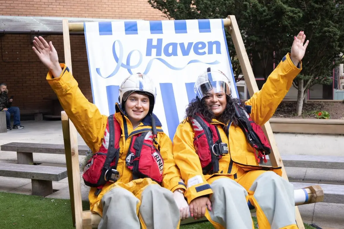 Two RNLI volunteers in yellow waterproofs and helmets wave while sitting in a large Haven deckchair.