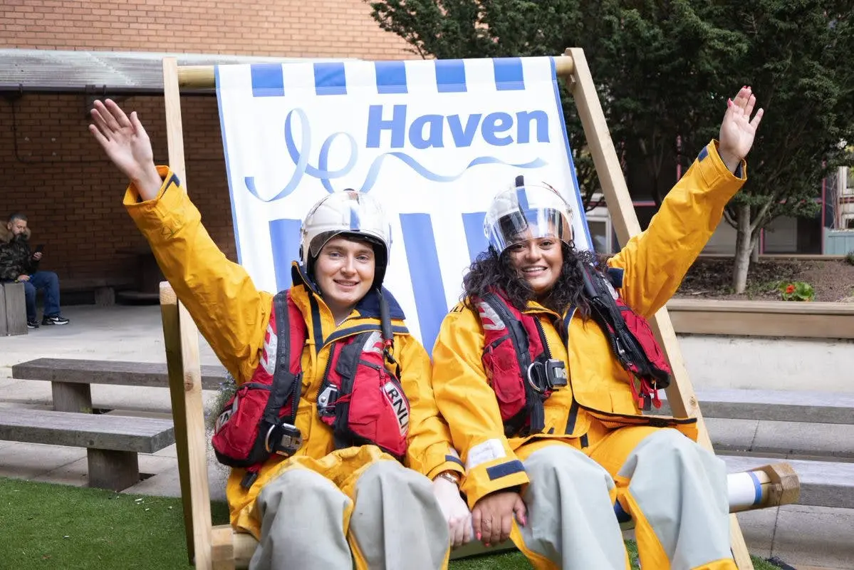 Two RNLI volunteers in yellow waterproofs and helmets wave while sitting in a large Haven deckchair.