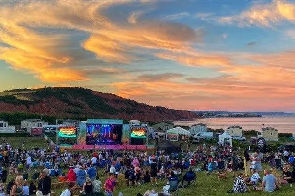 A large crowd gathers at an outdoor music festival by the coast during a vibrant sunset.