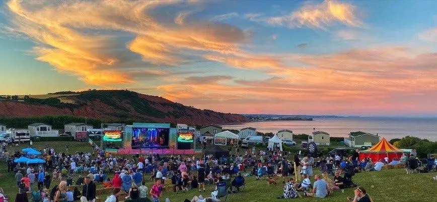 A large crowd gathers at an outdoor music festival by the coast during a vibrant sunset.