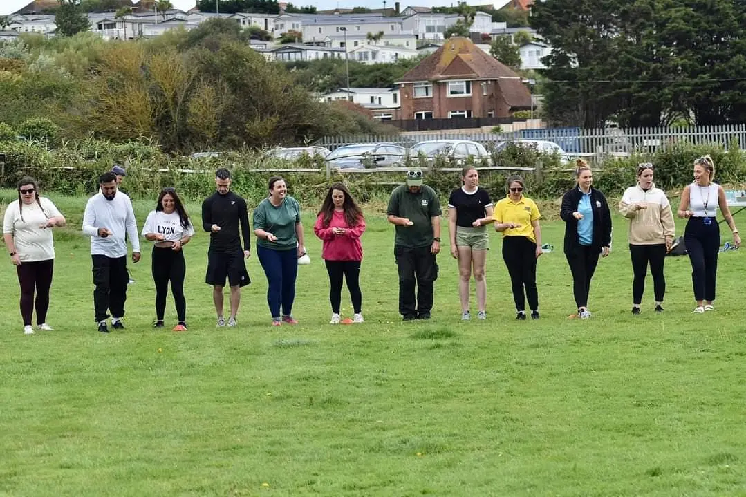 A group of adults lined up on a grassy field, participating in an egg and spoon race.