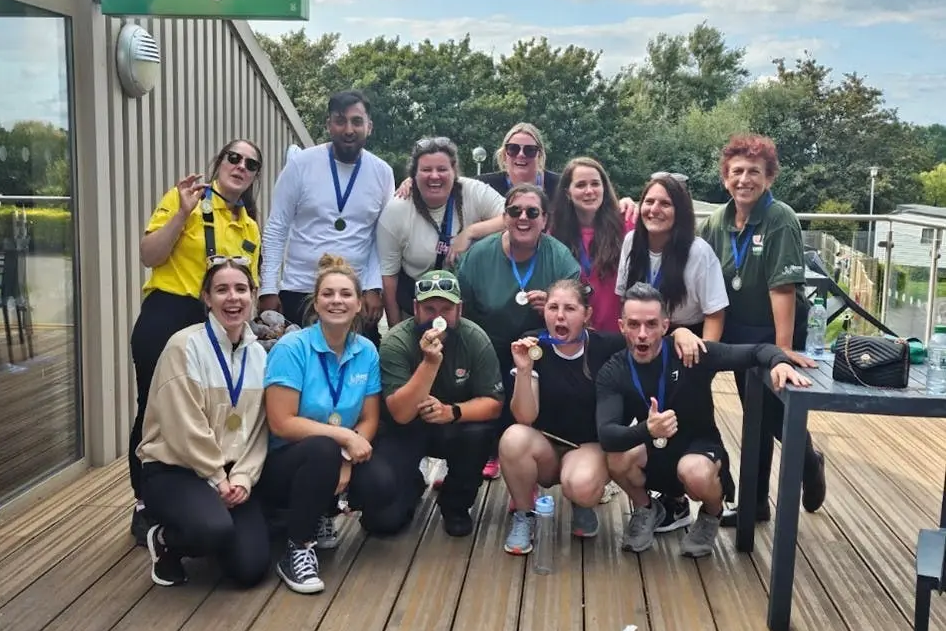 A large group of smiling adults pose together outdoors on a wooden deck, proudly wearing medals around their necks.