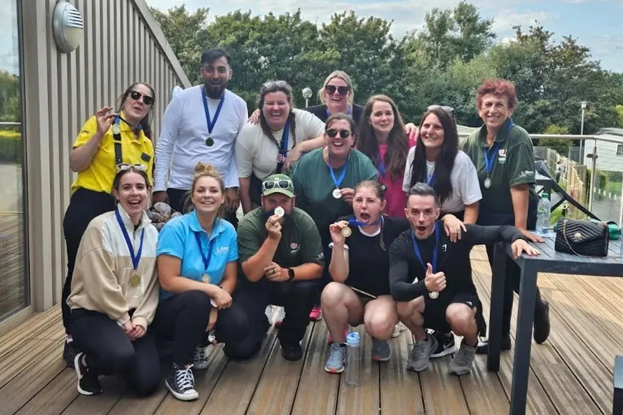 A large group of smiling adults pose together outdoors on a wooden deck, proudly wearing medals around their necks.