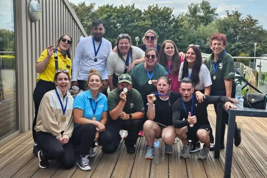 A large group of smiling adults pose together outdoors on a wooden deck, proudly wearing medals around their necks.