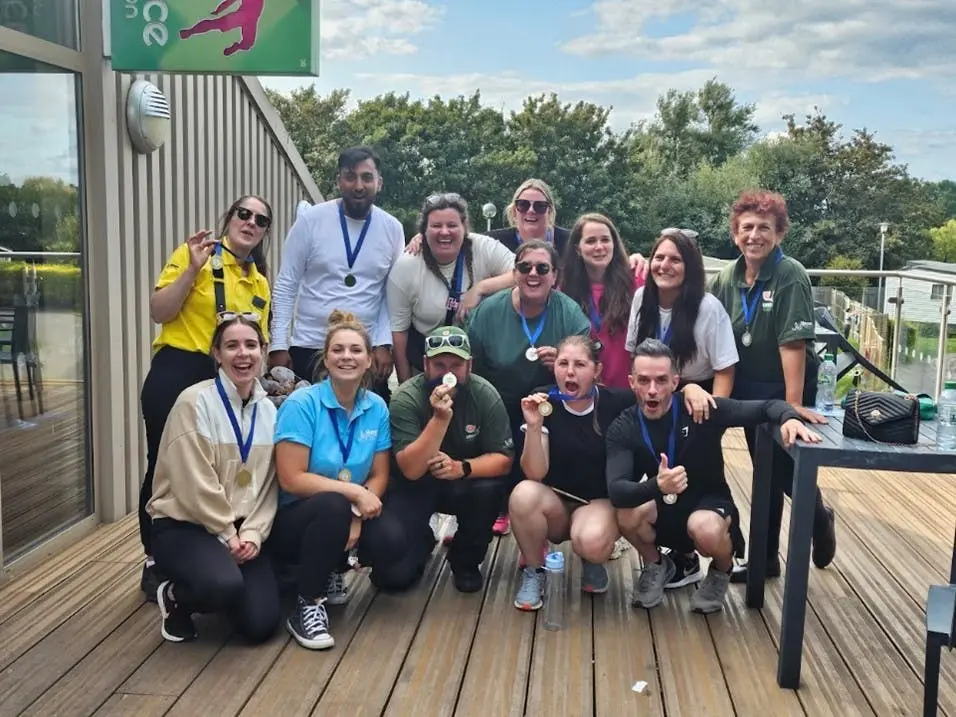 A large group of smiling adults pose together outdoors on a wooden deck, proudly wearing medals around their necks.