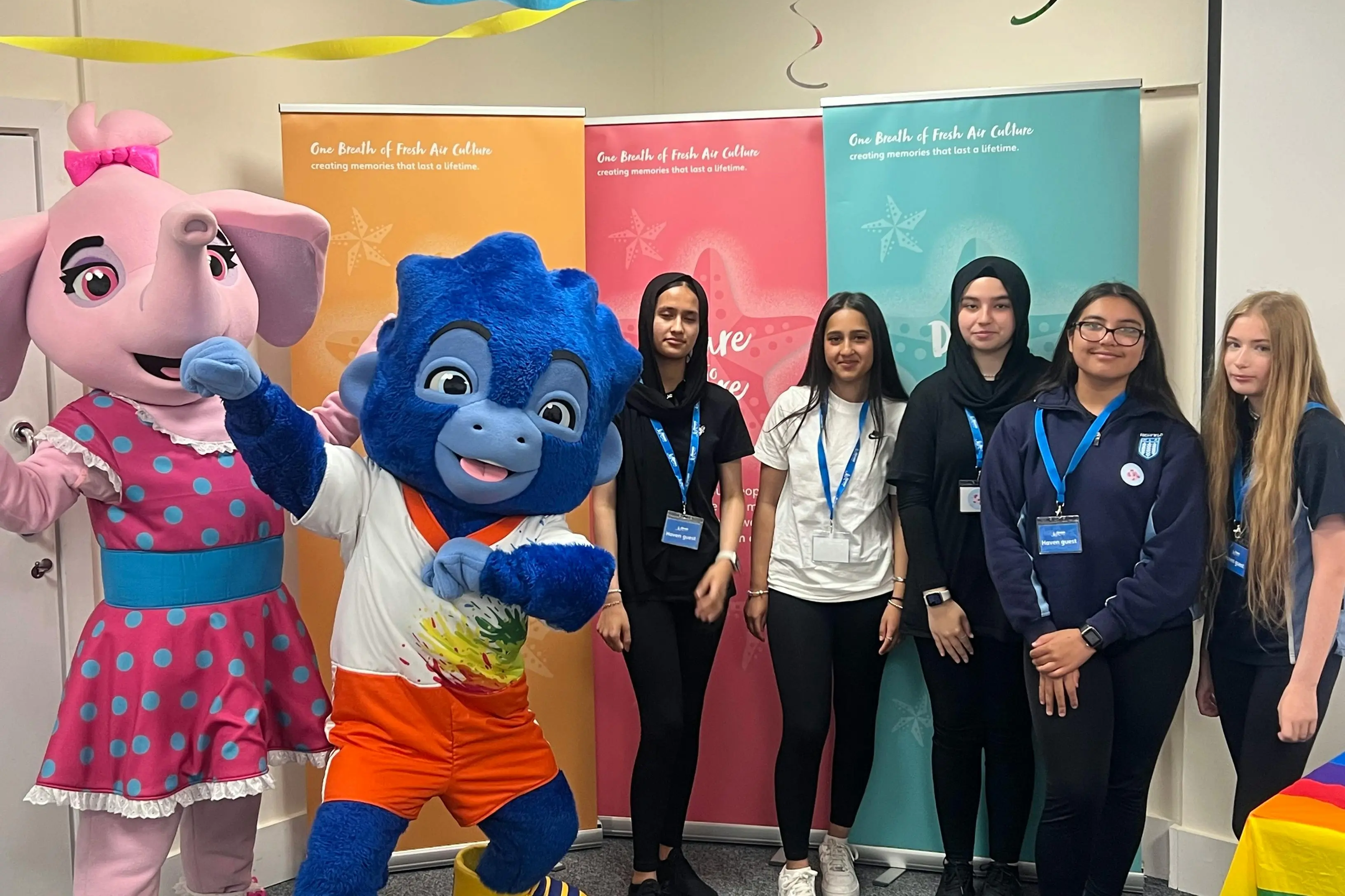 Pink elephant and blue monster mascots pose with five teenage girls wearing lanyards indoors.