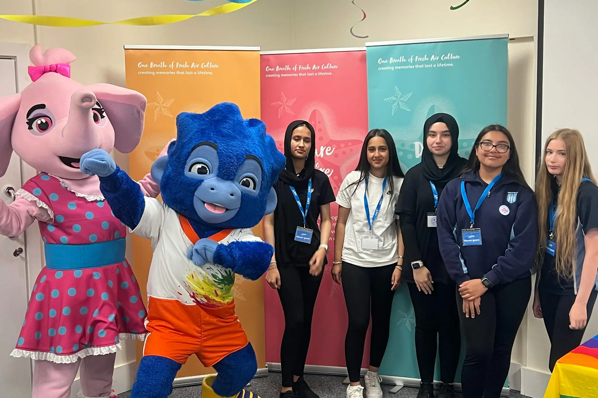 Group of young people posing with two colorful mascot costumes and event banners.