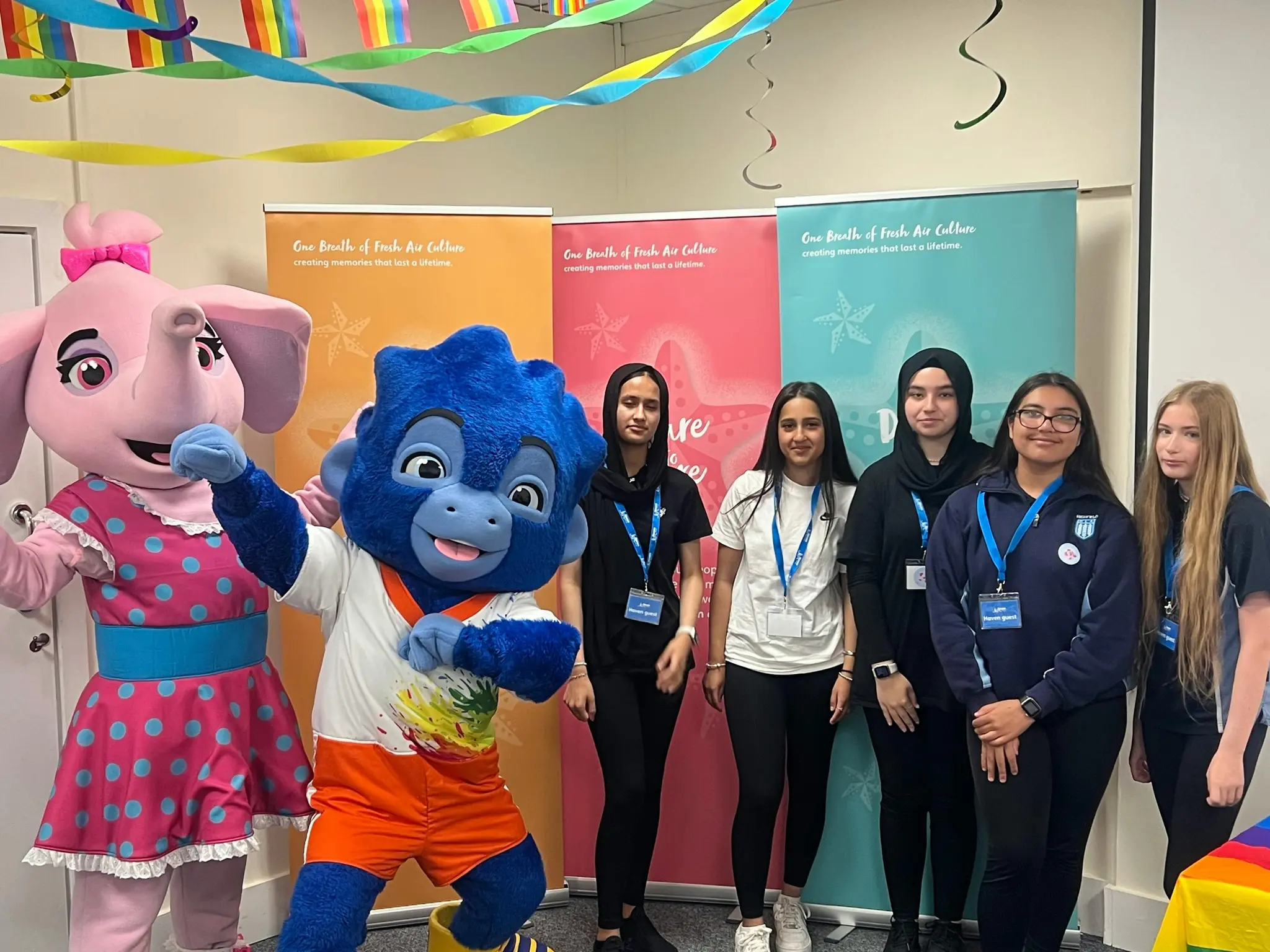 Group of young people posing with two colorful mascot costumes and event banners.