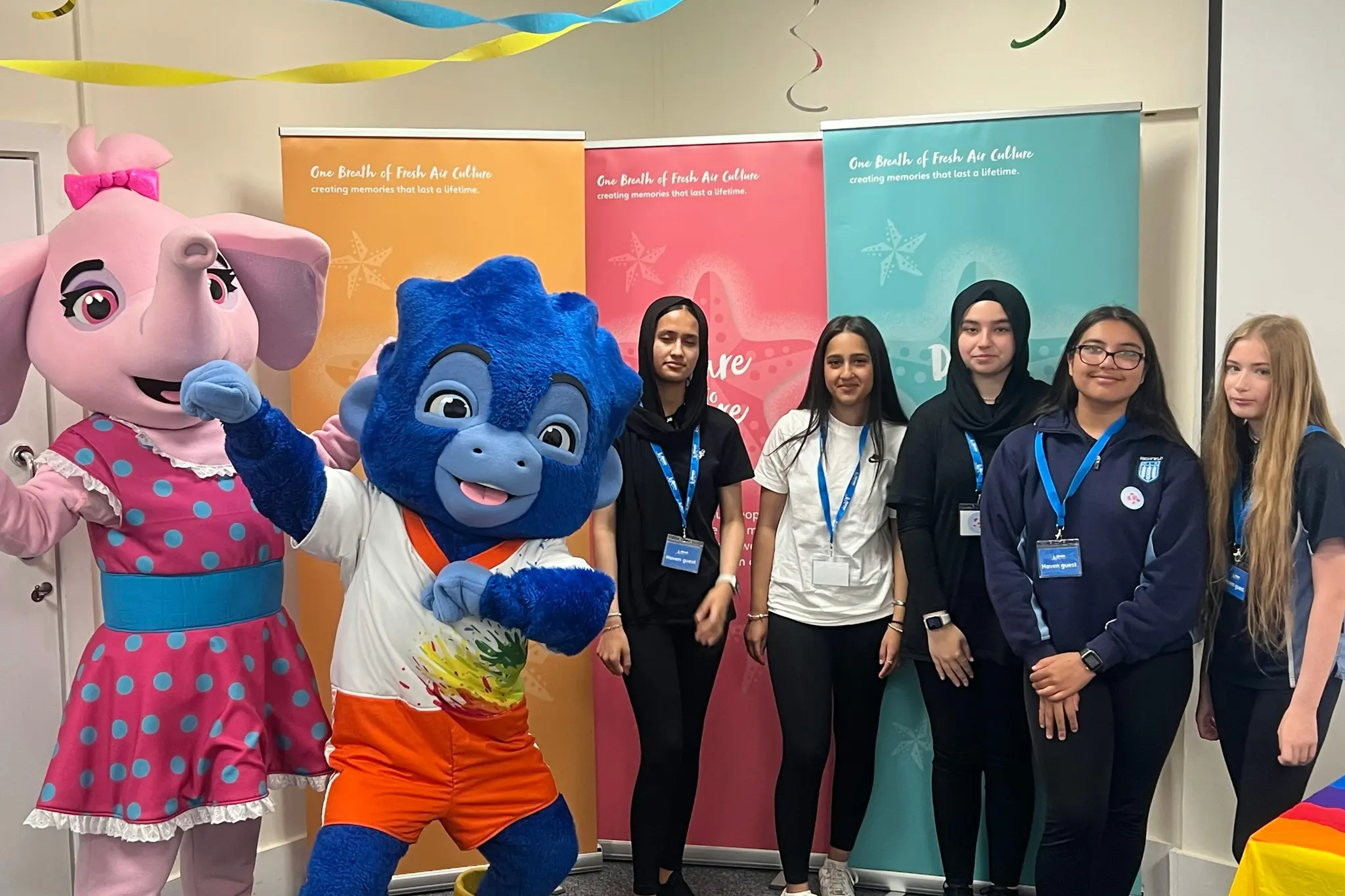 Group of young people posing with two colorful mascot costumes and event banners.
