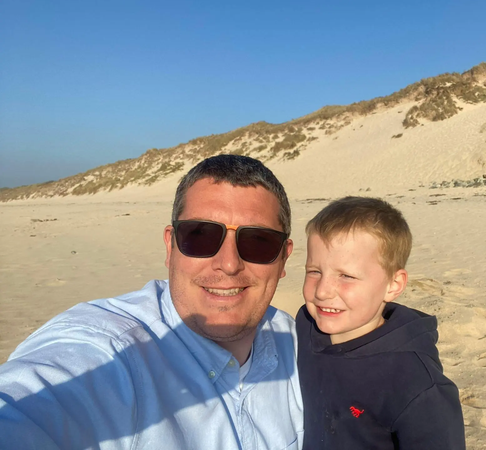 A father and son smiling together for a selfie on a sandy beach with dunes in the background.