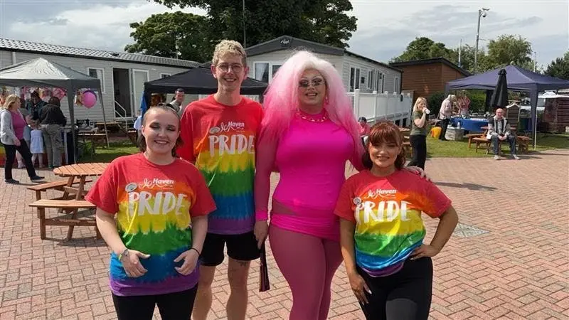 Four people posing outdoors in pride shirts, including one in a bright pink outfit.