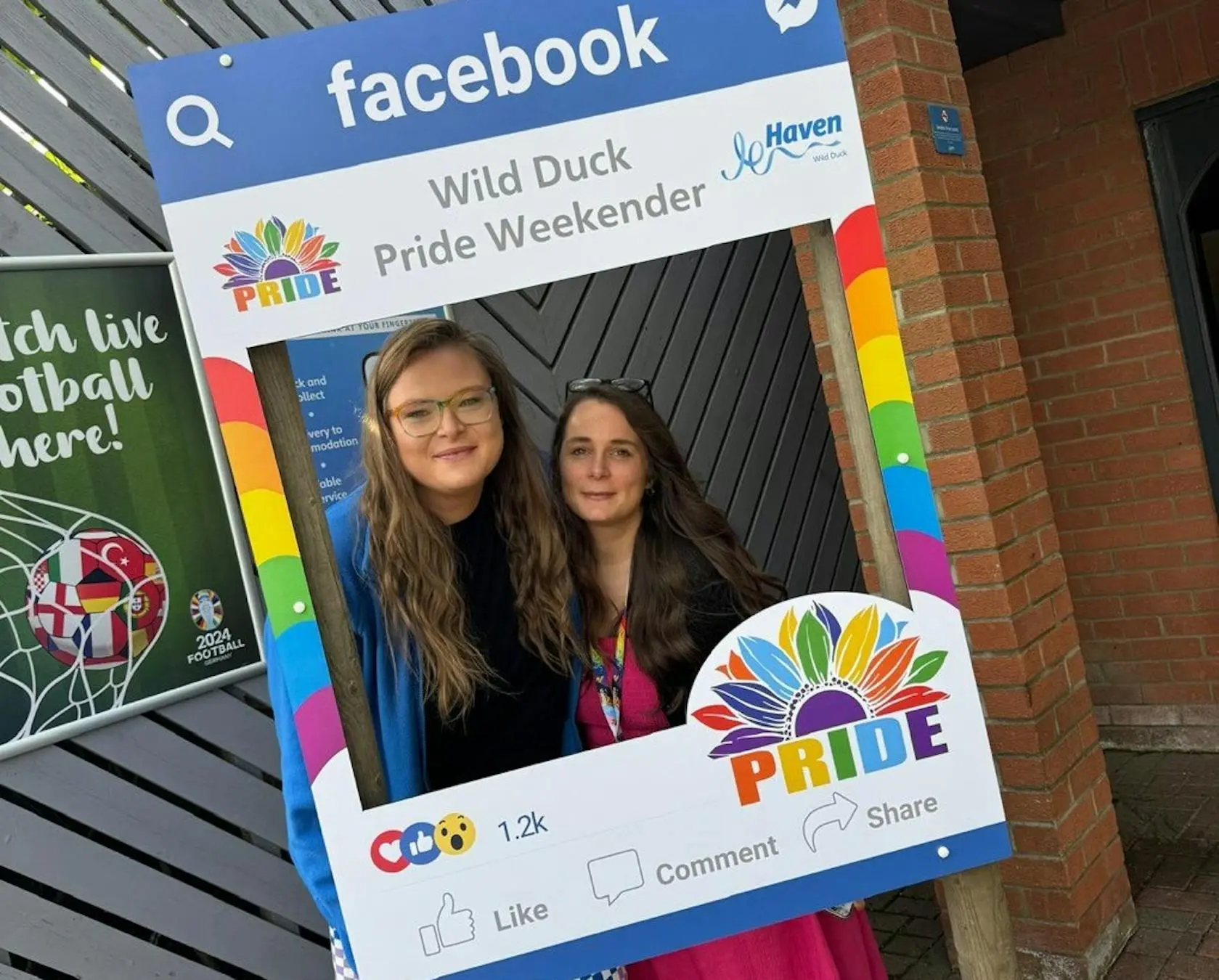Two women posing inside a Facebook-themed photo frame at the Wild Duck Pride Weekender.