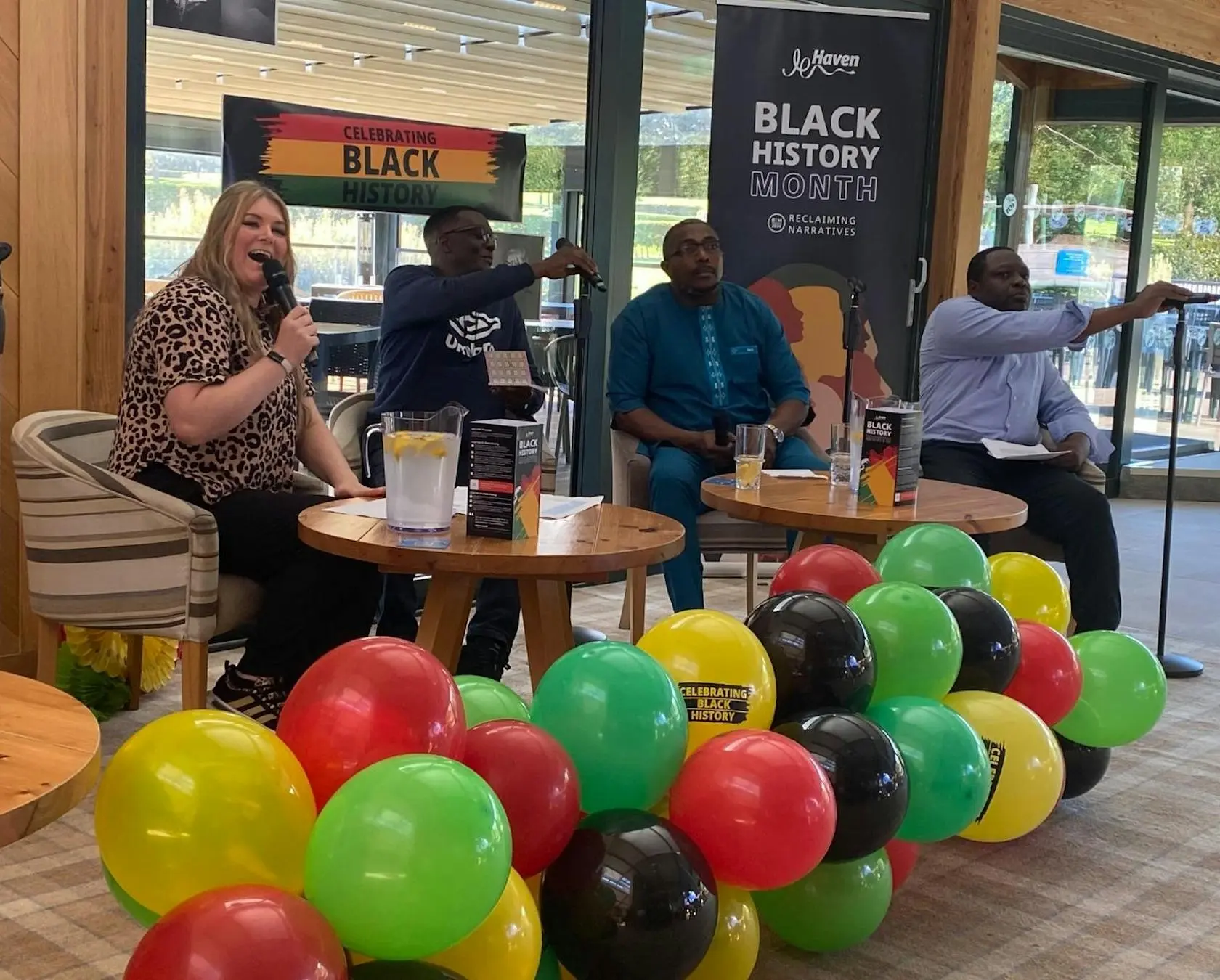 A panel of four people speaking at a Black History Month event with colorful balloons in the foreground.