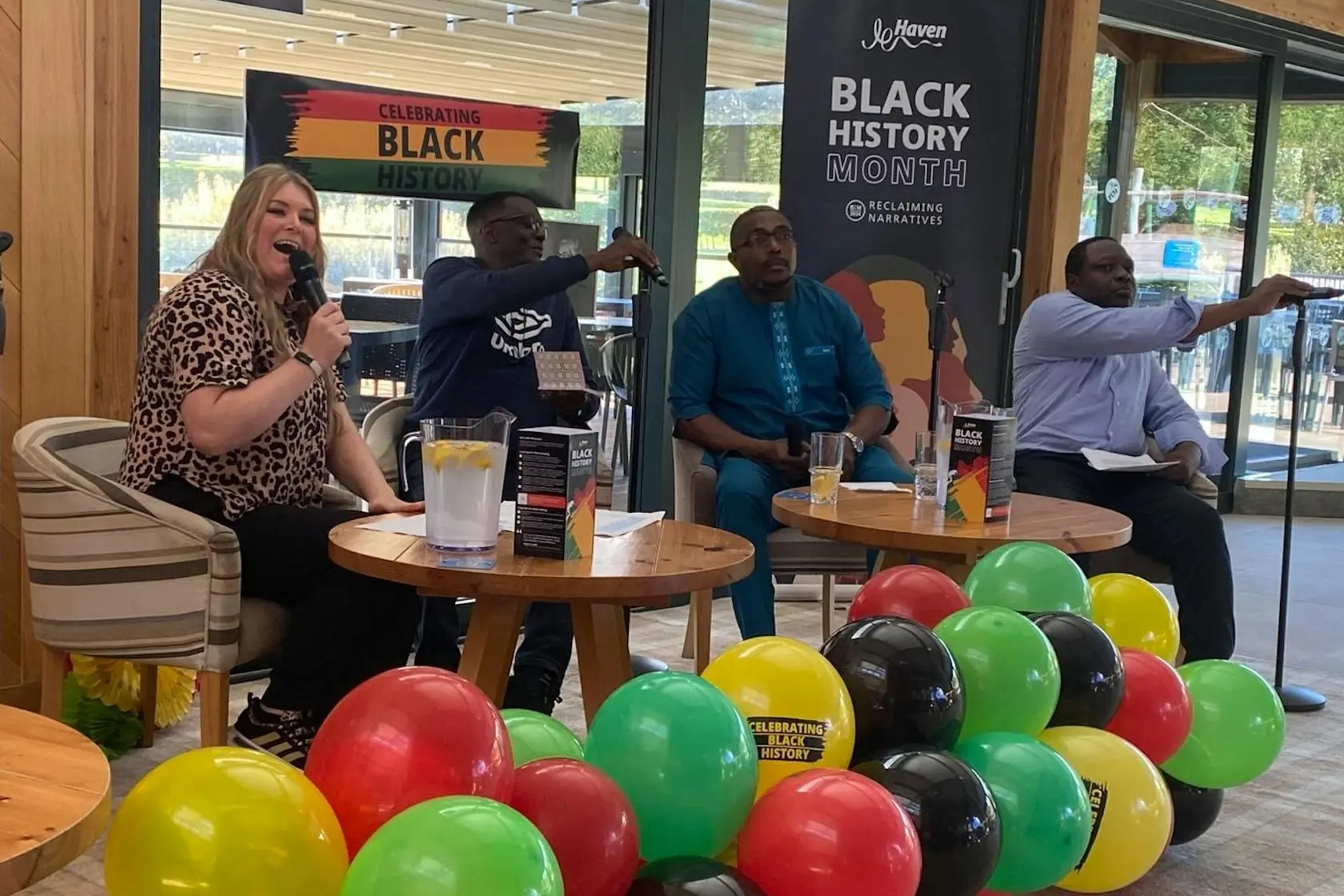 A panel of four people speaking at a Black History Month event with colorful balloons in the foreground.
