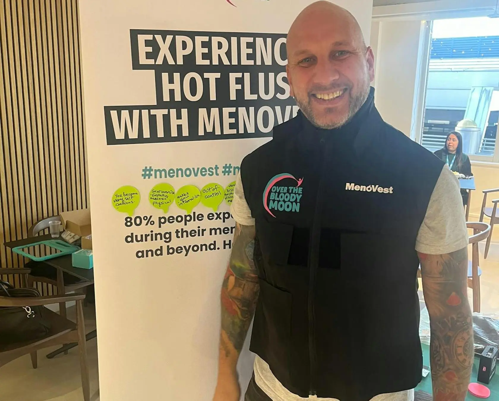 A smiling bald man wearing a MenoVest stands in front of an informational banner about hot flashes.