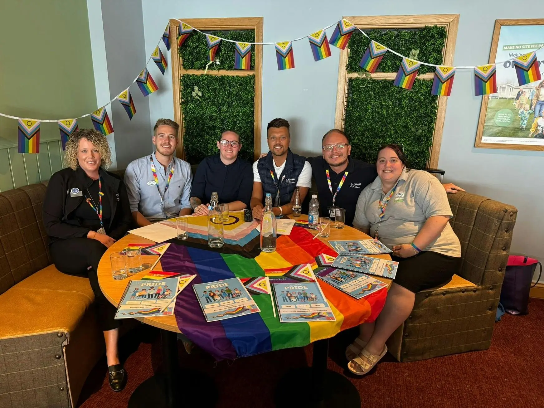 Six team members smiling around a table decorated with Pride flags and booklets, under Pride bunting.