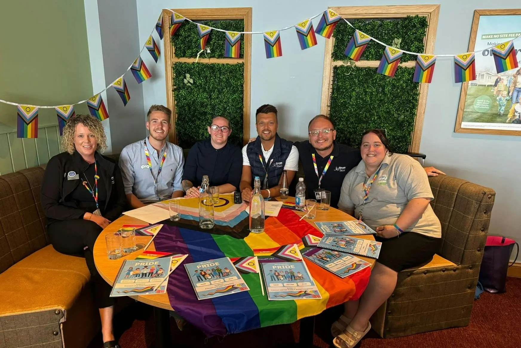 Six team members smiling around a table decorated with Pride flags and booklets, under Pride bunting.