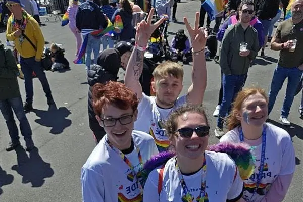 A group of friends smiling and taking a selfie at a crowded pride parade on a sunny day.