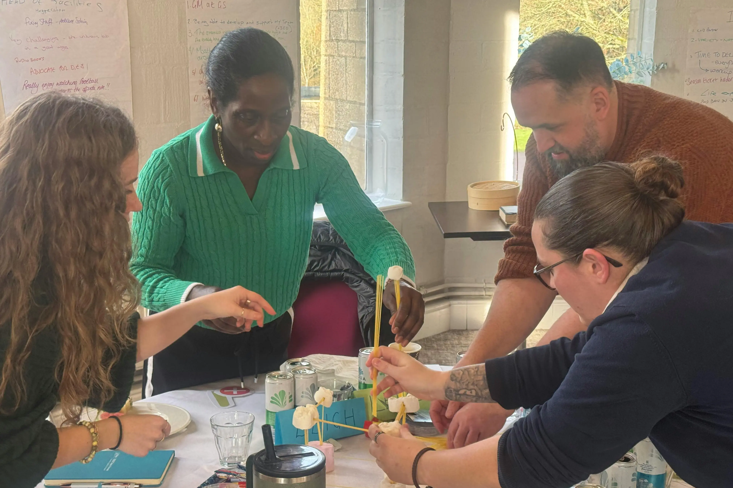 Four adults collaborating around a table to build a structure with marshmallows and spaghetti.