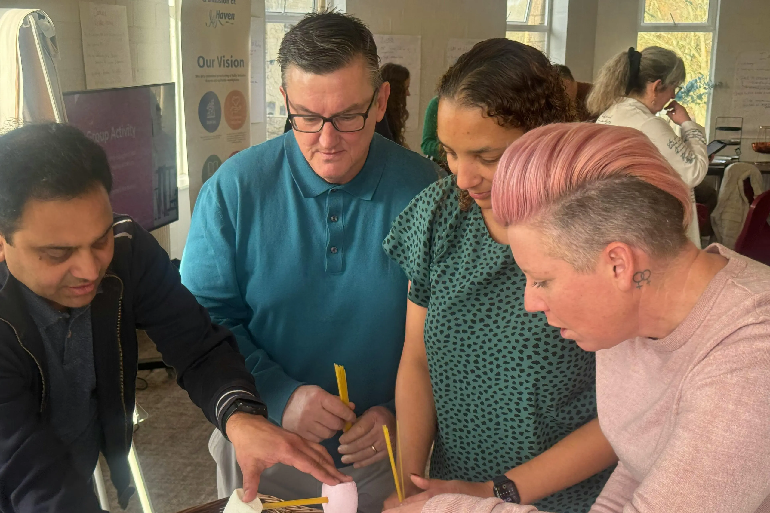 Four diverse colleagues collaborating on a marshmallow and spaghetti tower building exercise in an office.
