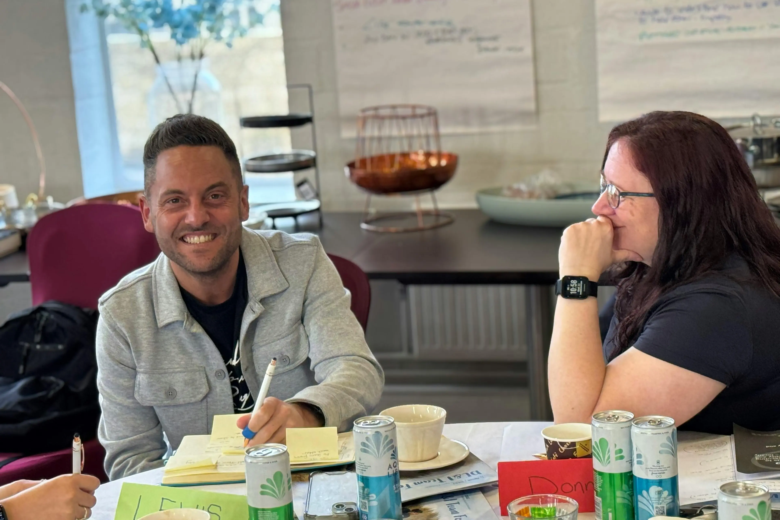 A man smiling at a table during a team workshop with a woman sitting next to him.