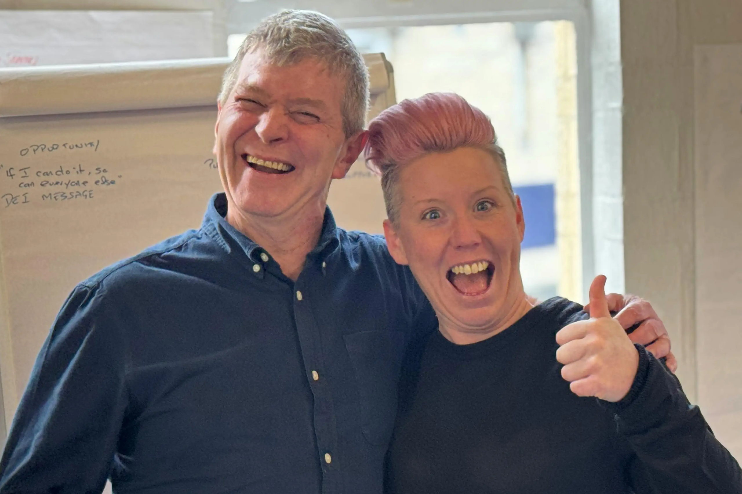 Two smiling colleagues standing next to each other, one giving a thumbs-up gesture.