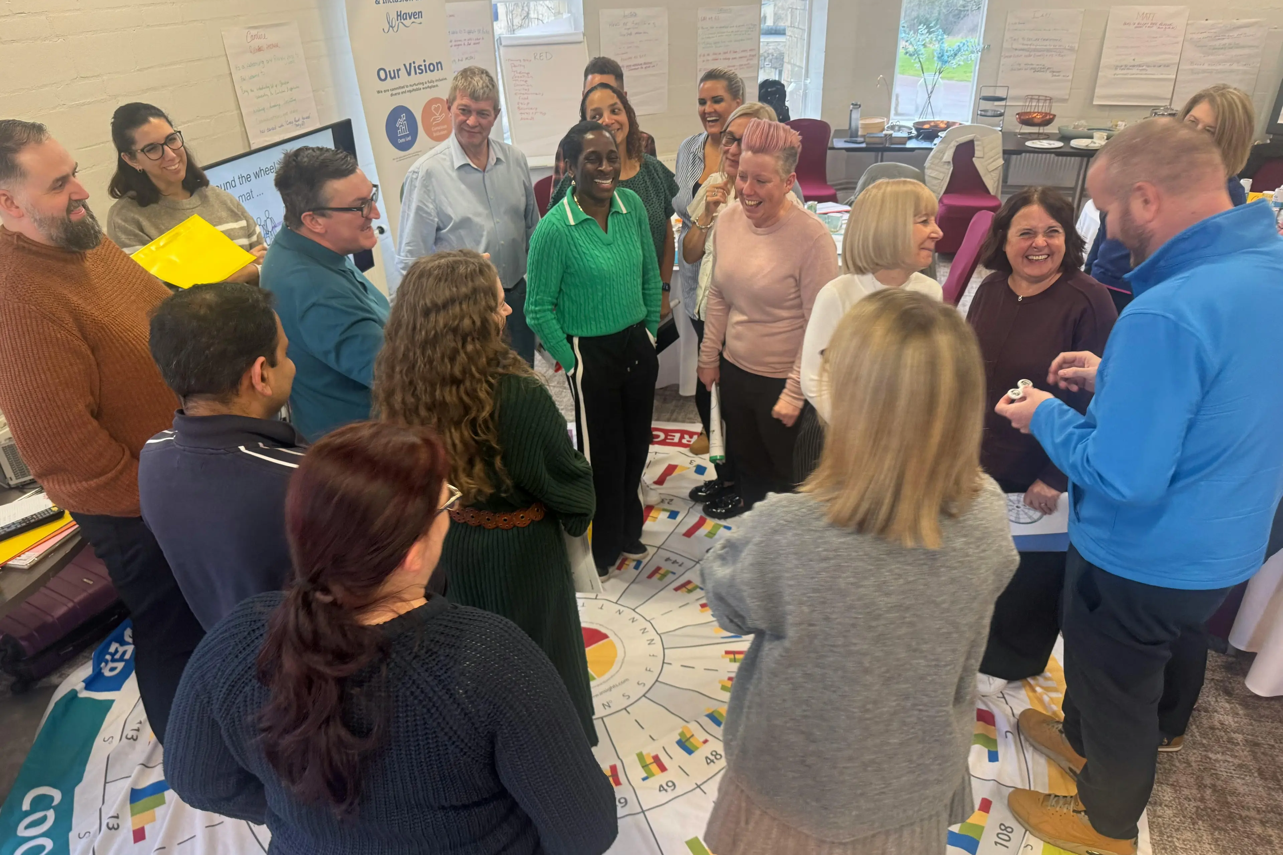 A diverse group of people gather in a circle around a large floor graphic during a workshop session.