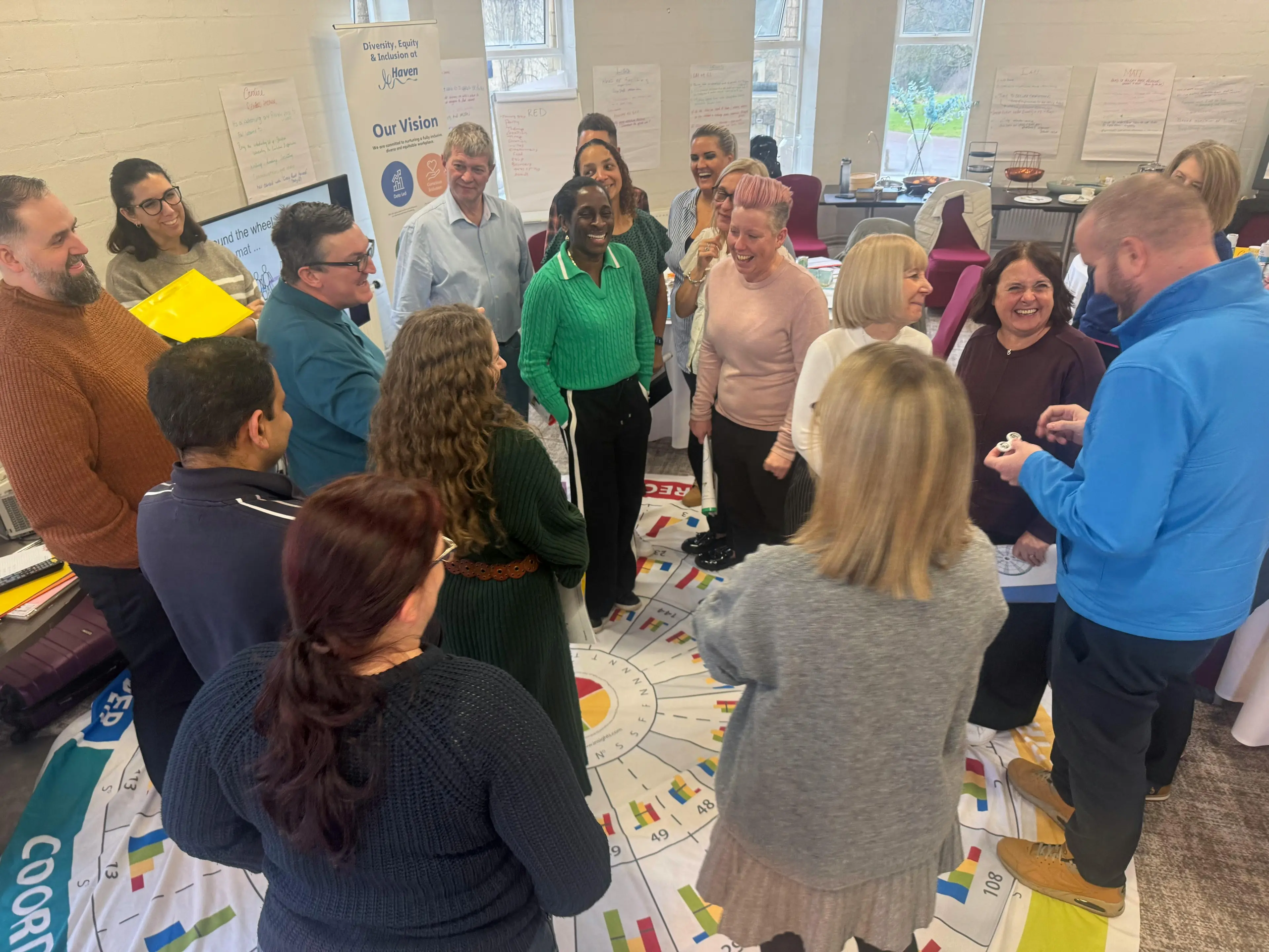 A diverse group of people gather in a circle around a large floor graphic during a workshop session.