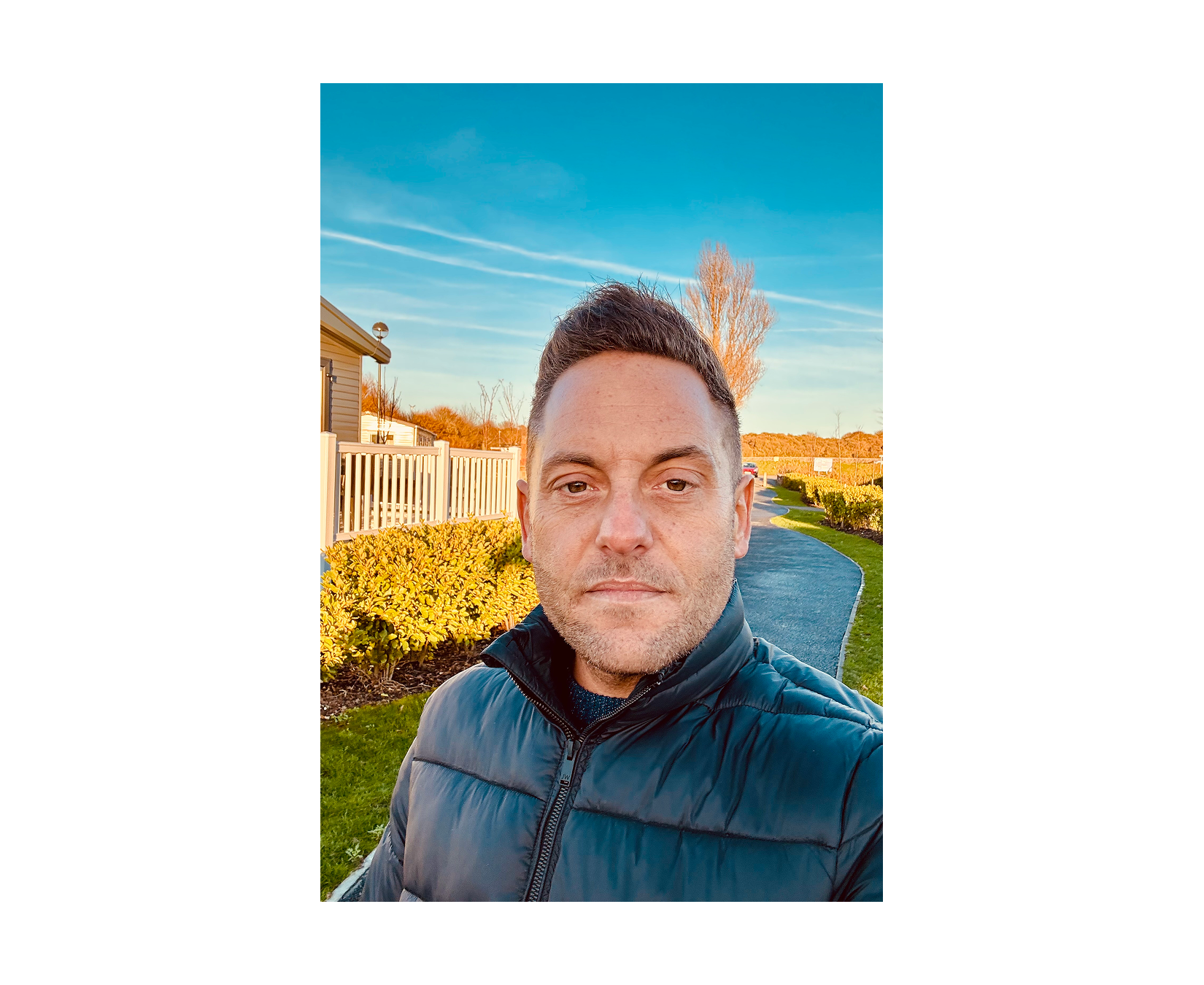 A man wearing a dark puffer jacket stands outside on a sunny day with a blue sky and path behind him.