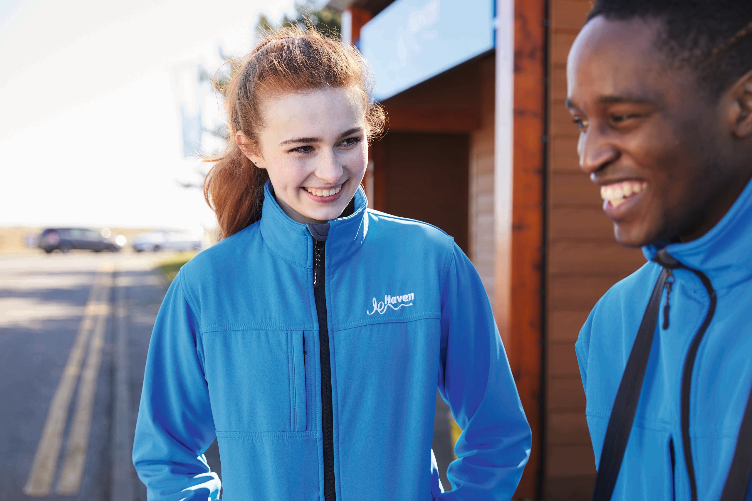 Two smiling colleagues wearing blue Haven jackets outdoors, interacting cheerfully.