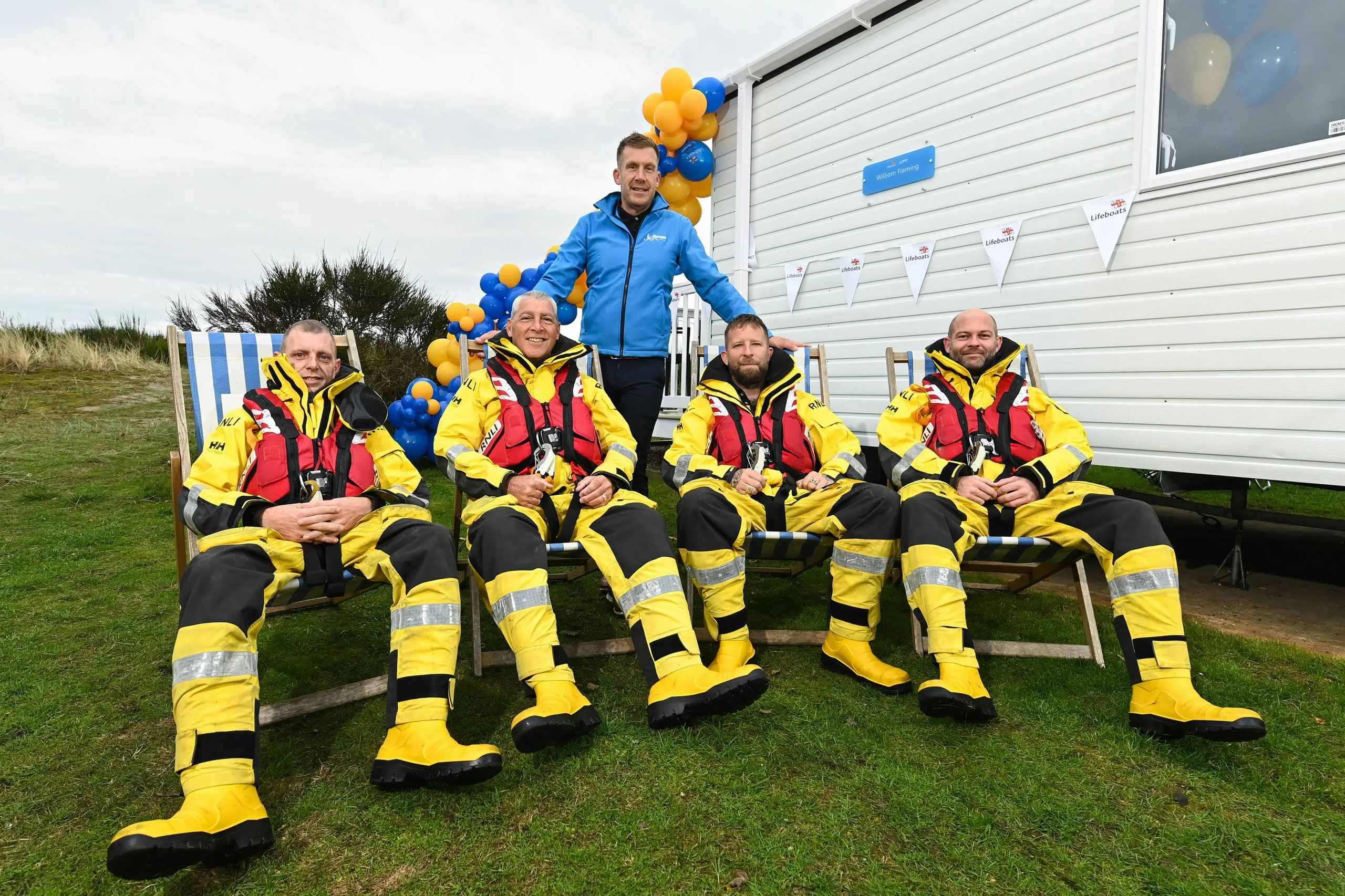 Four crew members in yellow RNLI waterproof suits sitting in deckchairs outside a white building.