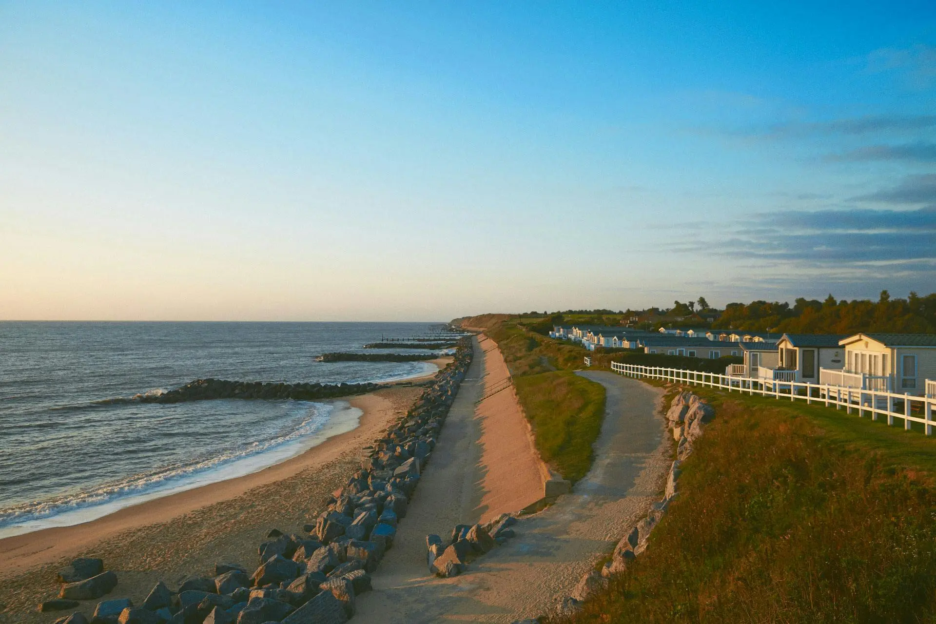A scenic view of a sandy beach, rocky breakwaters, and a row of coastal houses under a clear sky.