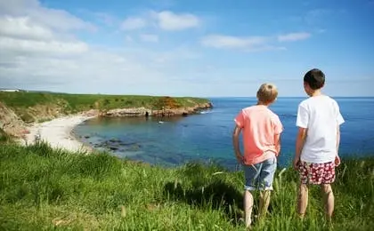 Two young boys standing on a grassy cliff overlooking a scenic ocean bay with a small sandy beach.