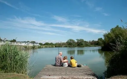 A grandfather and two children sitting on a wooden pier by a lake, fishing on a sunny day.