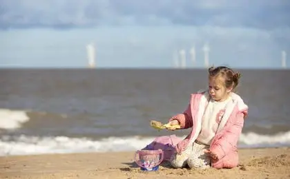 A young girl in a pink jacket plays with a bucket and spade on a sandy beach by the sea.