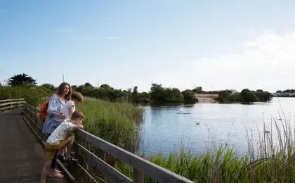 A woman and two children stand on a wooden boardwalk, looking out over a calm wetland lake on a sunny day.